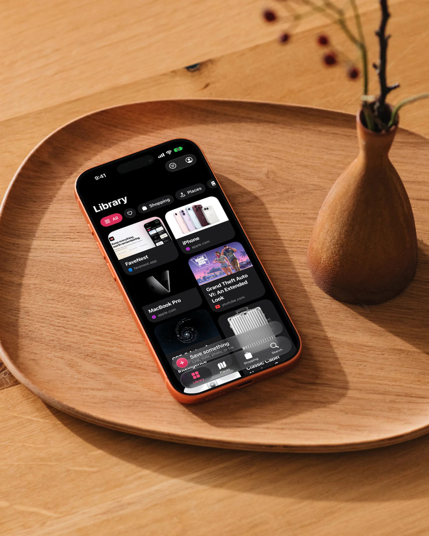 An iPhone lying on a wooden serving tray beside a small wooden vase, showing the FaveNest Library in dark mode: a grid of saved cards including the FaveNest website, the iPhone and MacBook Pro pages from apple.com, and a Grand Theft Auto VI trailer from YouTube.